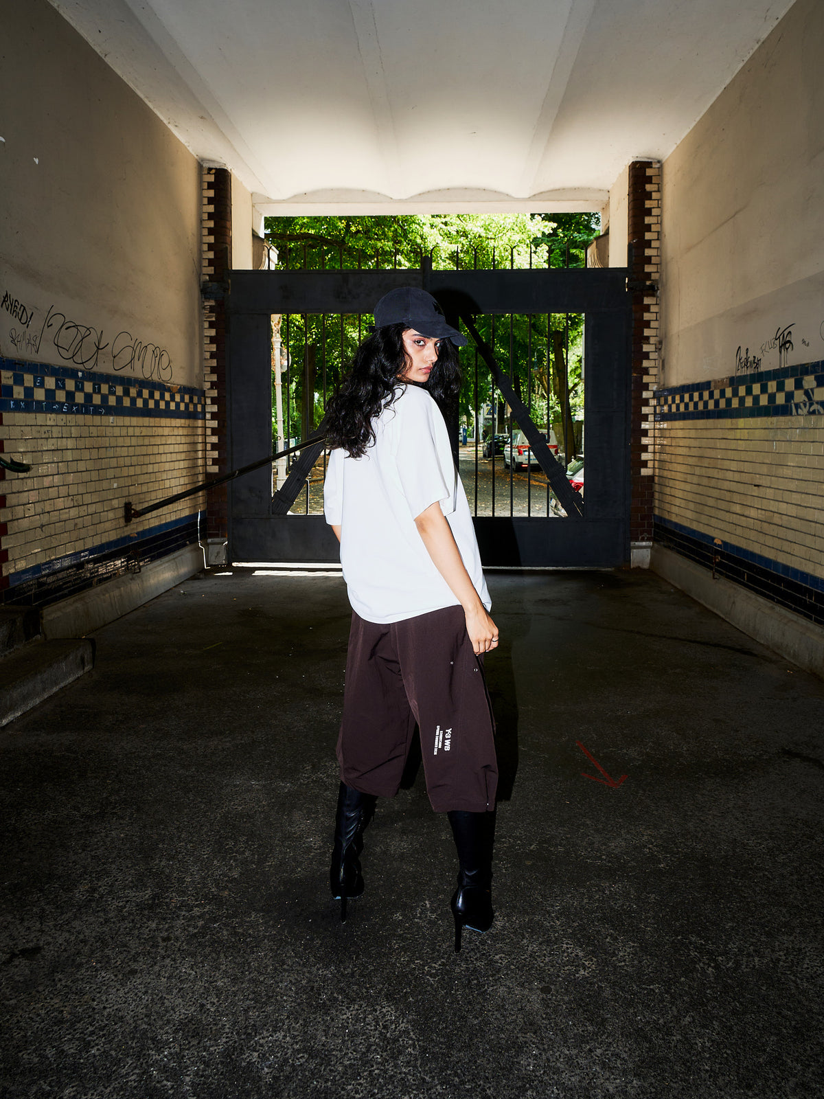 A model stands in a concrete tunnel wearing an oversized white t-shirt, dark brown cargo pants, black knee-high boots, and a black baseball cap, looking back over their shoulder.
