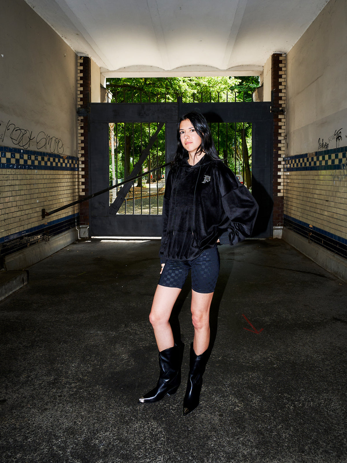 A model stands in a dark, industrial tunnel or underpass wearing a black fuzzy cropped jacket, black patterned shorts, and black pointed-toe boots. The background features a large black gate opening to a green forest, with brick walls on either side.