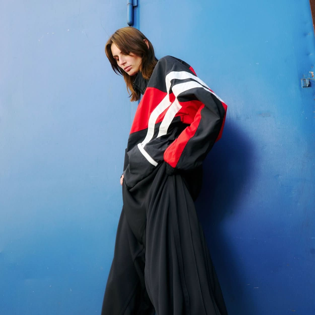 Young woman in front of blue background wearing a track jacket in red and black with shite stripes by VETEMENTS and wide black pants
