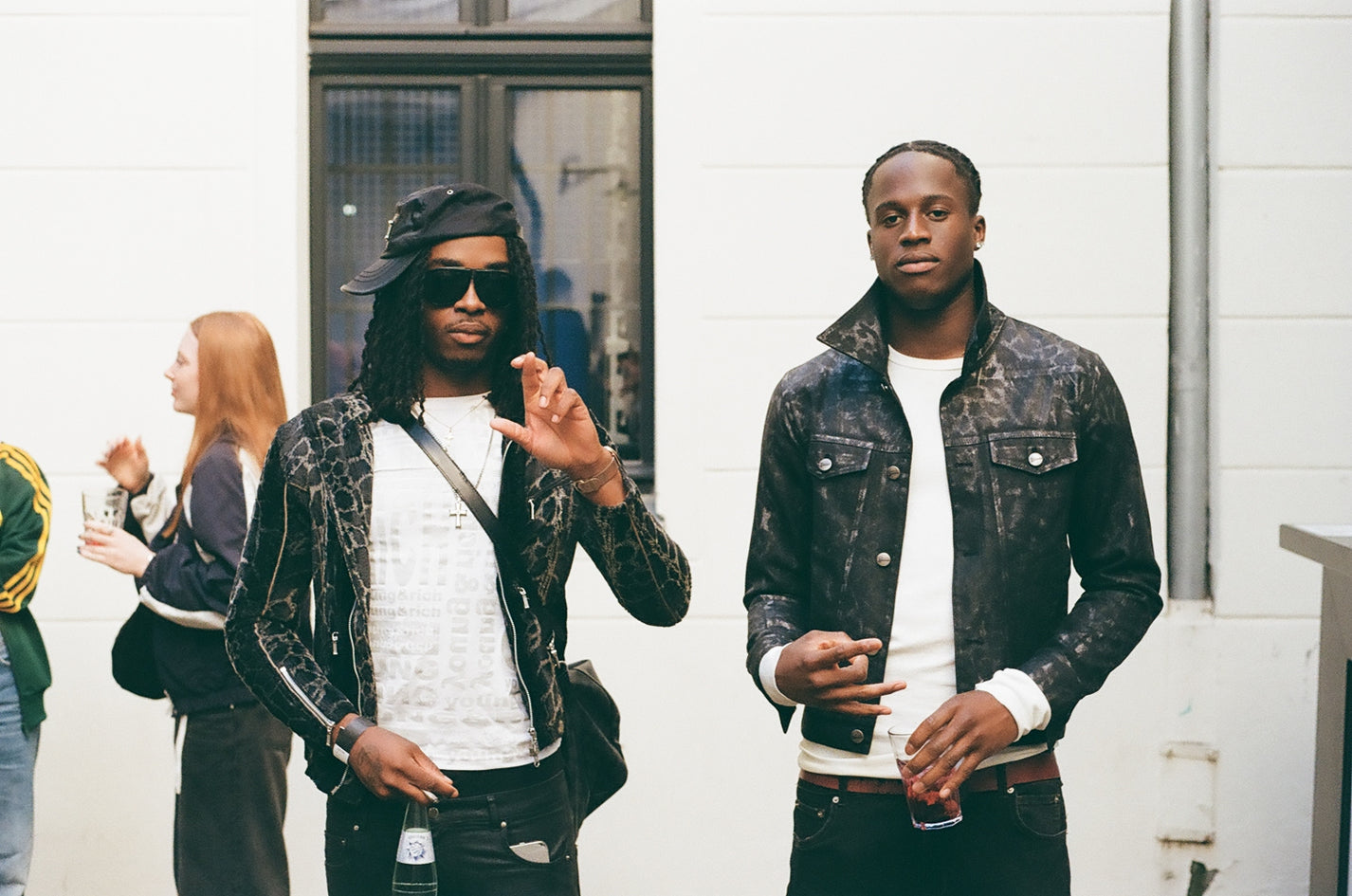 Puma x Non Faith Studios Event Recap. Two men standing outdoors, one making a hand gesture, with a white wall and glass door in the background.