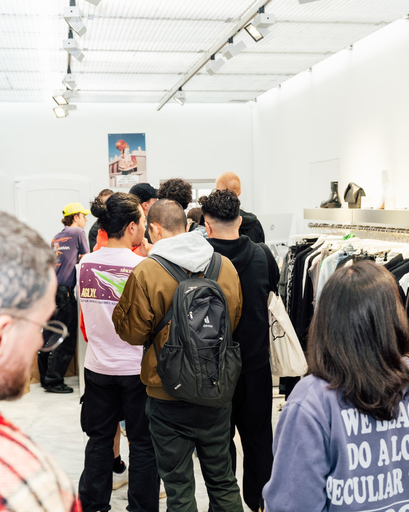 RECAP: Tom Sachs x Nike Mars Yard 3.0. People shopping in a clothing store with racks of clothes and a white wall in the background.