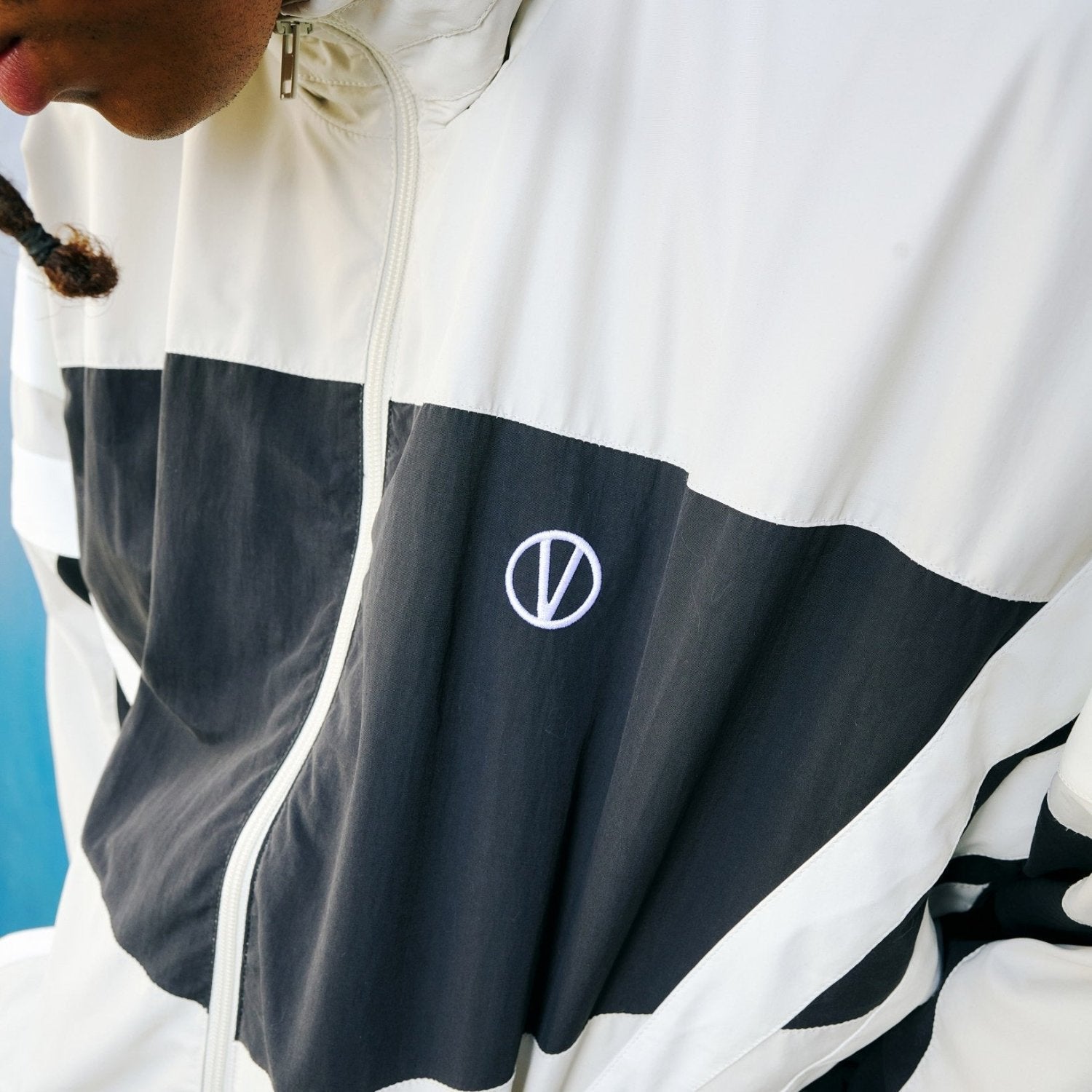 Close up image of a young man wearing a track jacket in beige and black with white stripes by VETEMENTS and wide black pants, focussing on the V detail on the chest