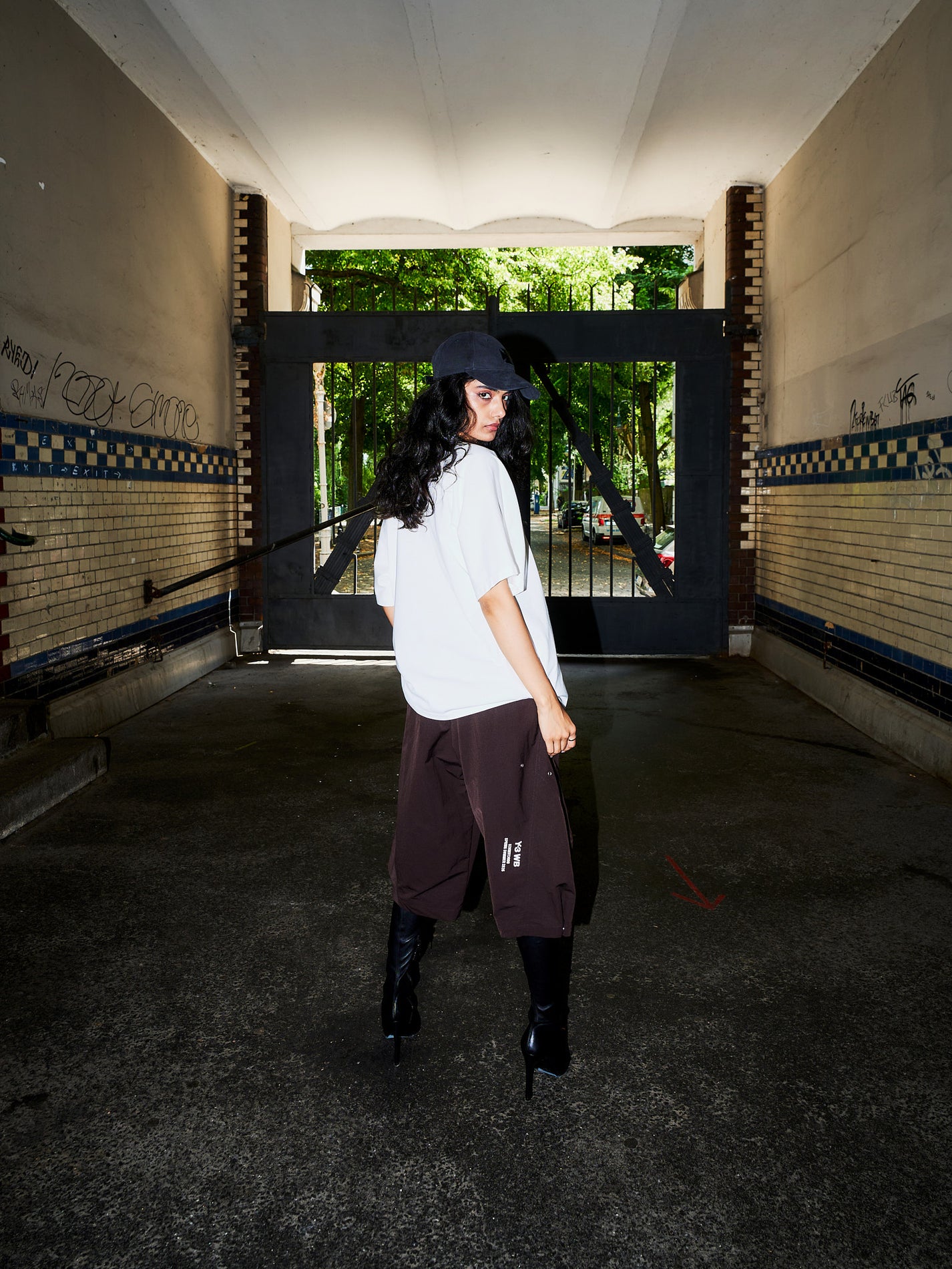 A model stands in a concrete tunnel wearing an oversized white t-shirt, dark brown cargo pants, black knee-high boots, and a black baseball cap, looking back over their shoulder.