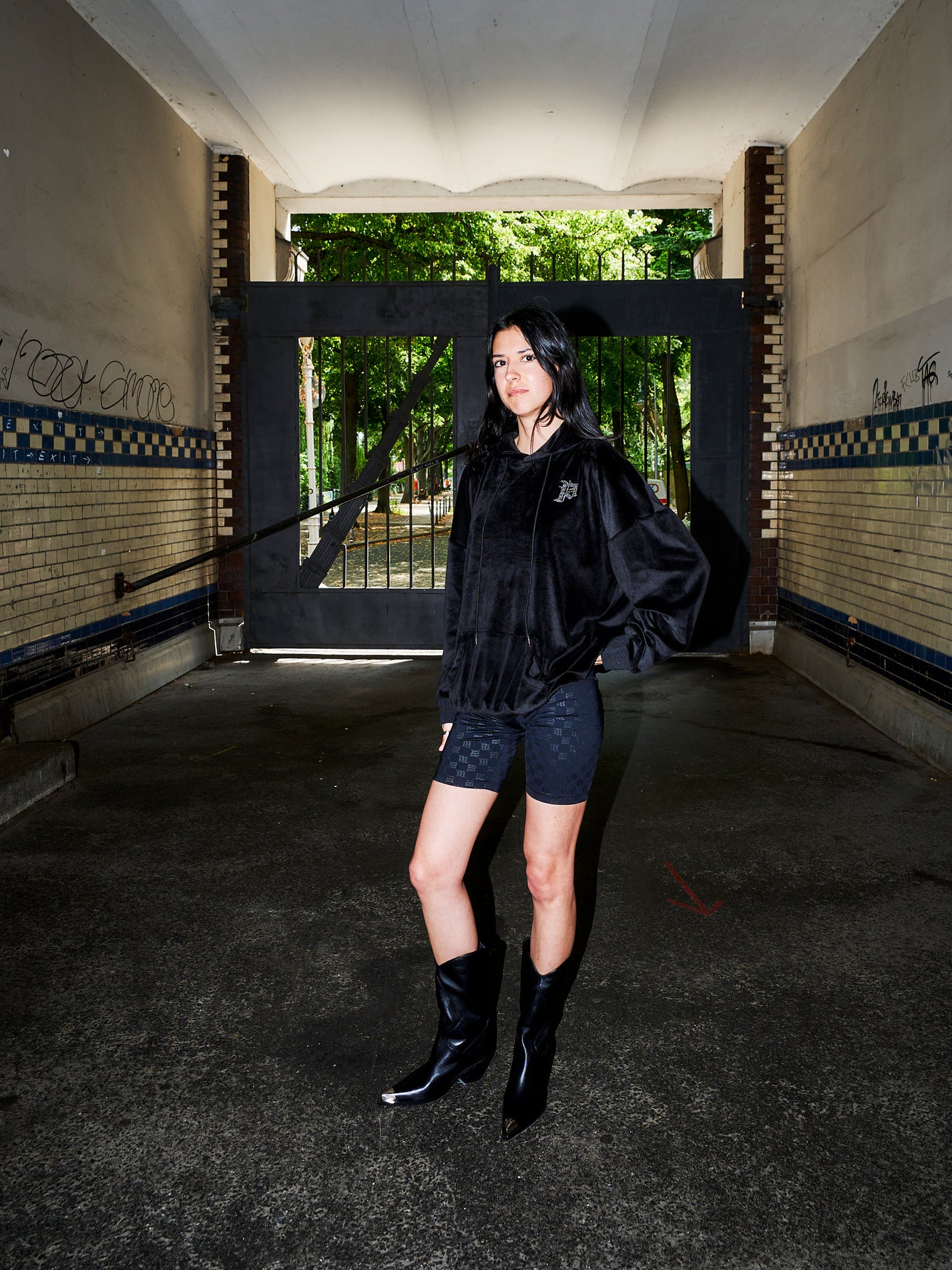 A model stands in a dark, industrial tunnel or underpass wearing a black fuzzy cropped jacket, black patterned shorts, and black pointed-toe boots. The background features a large black gate opening to a green forest, with brick walls on either side.