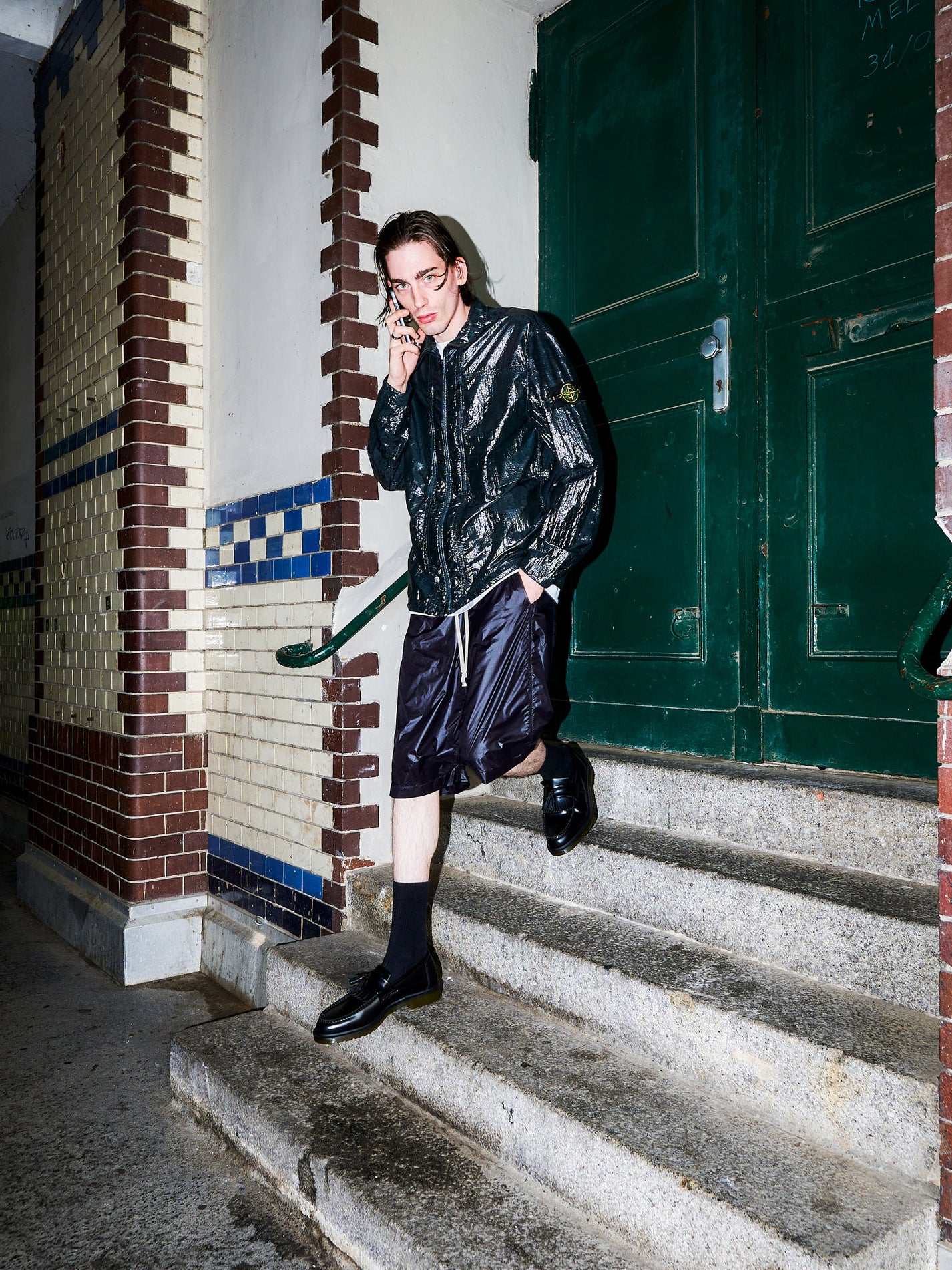 A model wearing a shiny black bomber jacket, matching black shorts, black socks, and black leather shoes walks down concrete stairs next to a green door and tiled wall.
