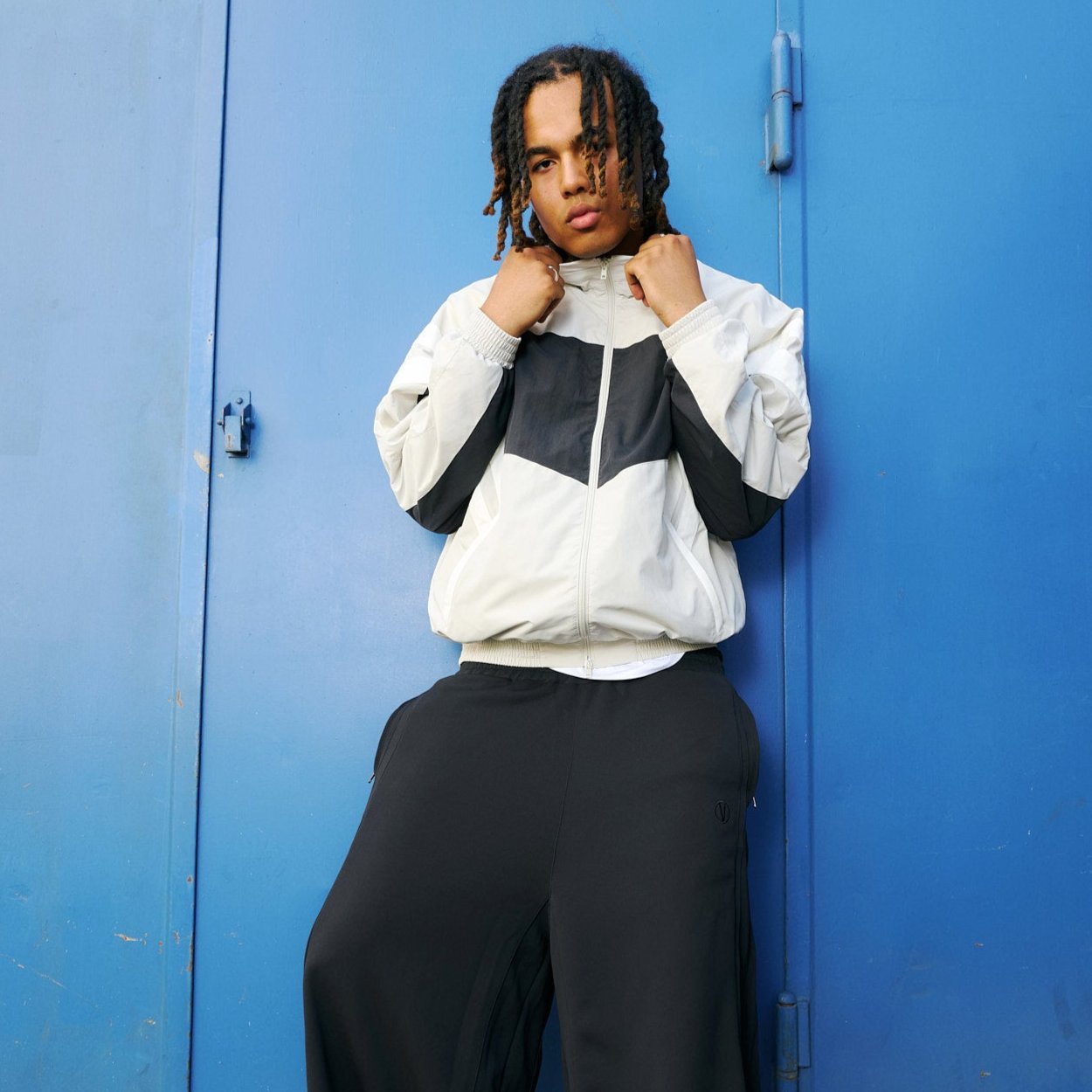 Young man  in front of blue background wearing a track jacket in beige and black with white stripes by VETEMENTS and wide black pants