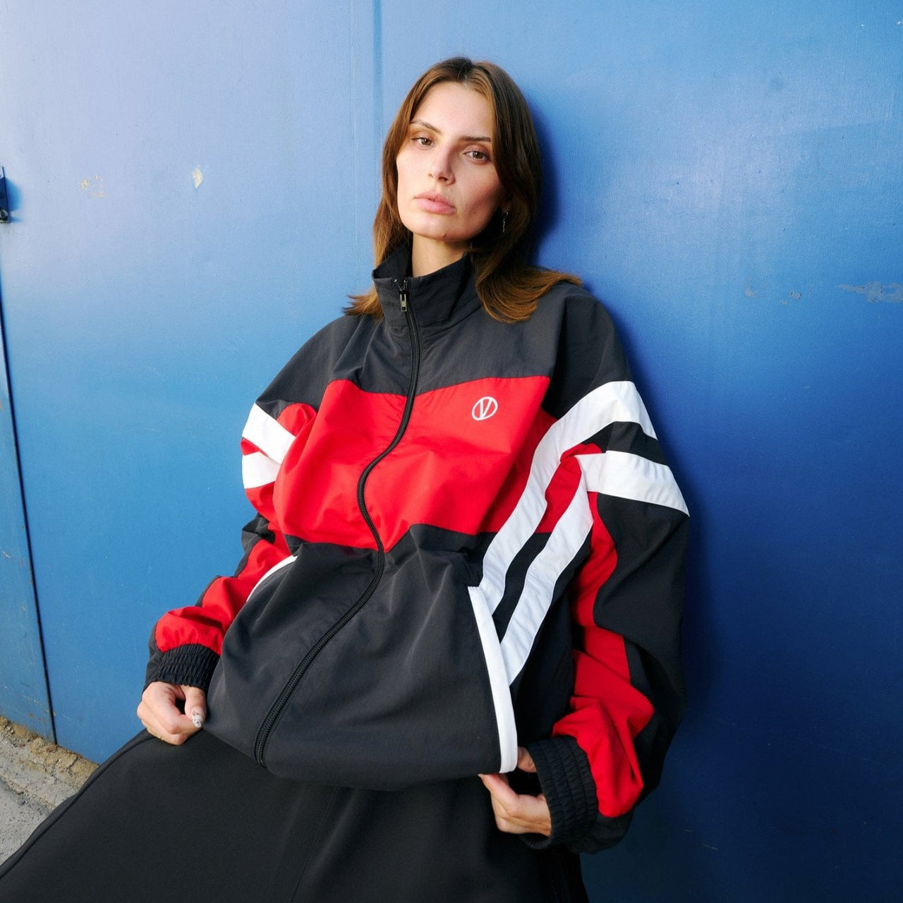 Young woman  in front of blue background wearing a track jacket in red and black with shite stripes by VETEMENTS