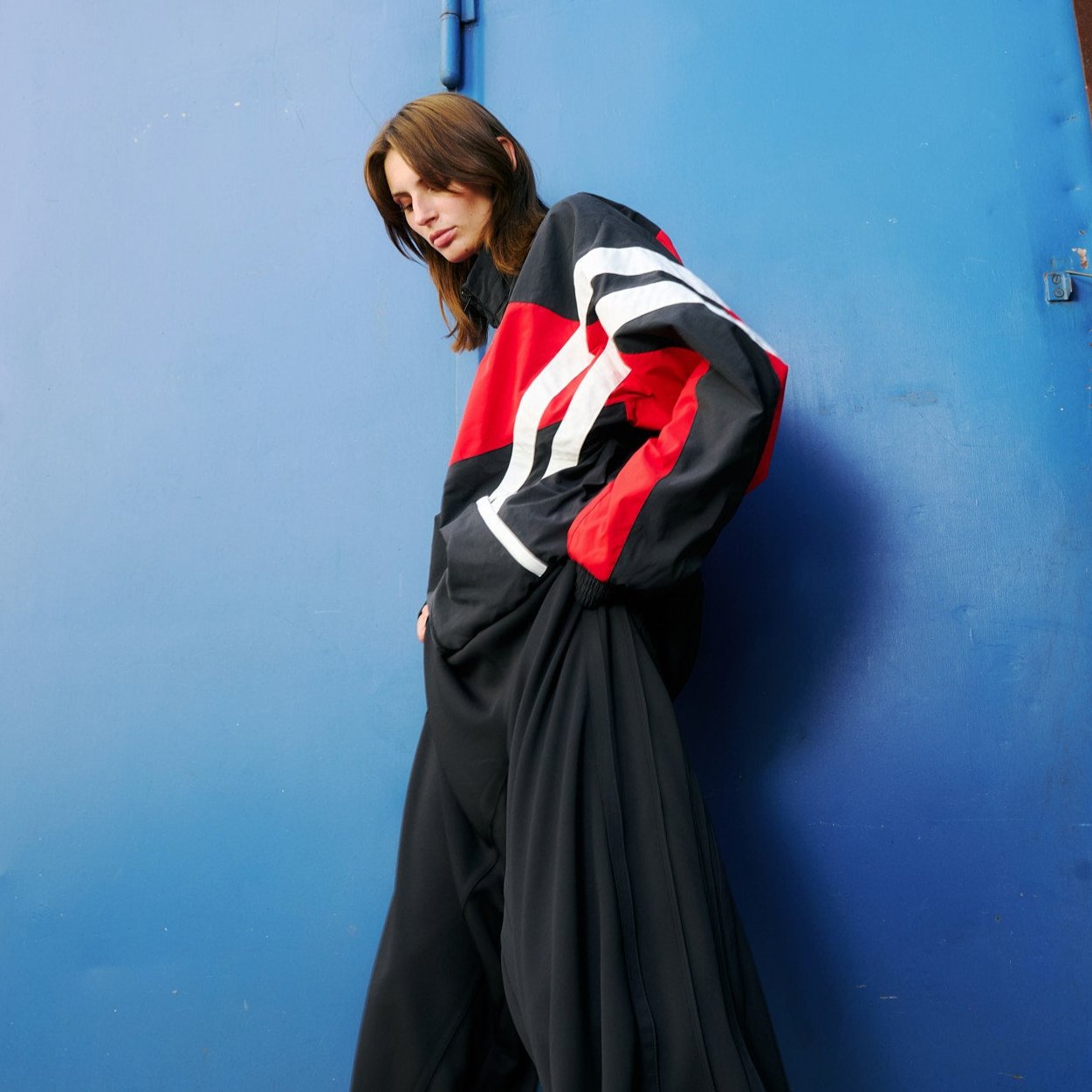 Young woman  in front of blue background wearing a track jacket in red and black with shite stripes by VETEMENTS