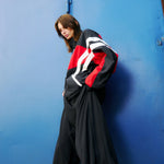 Young woman  in front of blue background wearing a track jacket in red and black with shite stripes by VETEMENTS and wide black pants