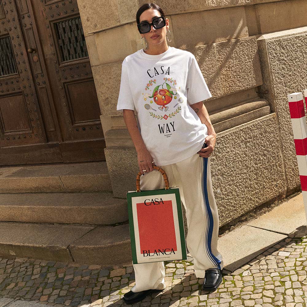 Person wearing a white t-shirt with 'Casa Way' design, holding a Casablanca bag, standing on a stone pavement.