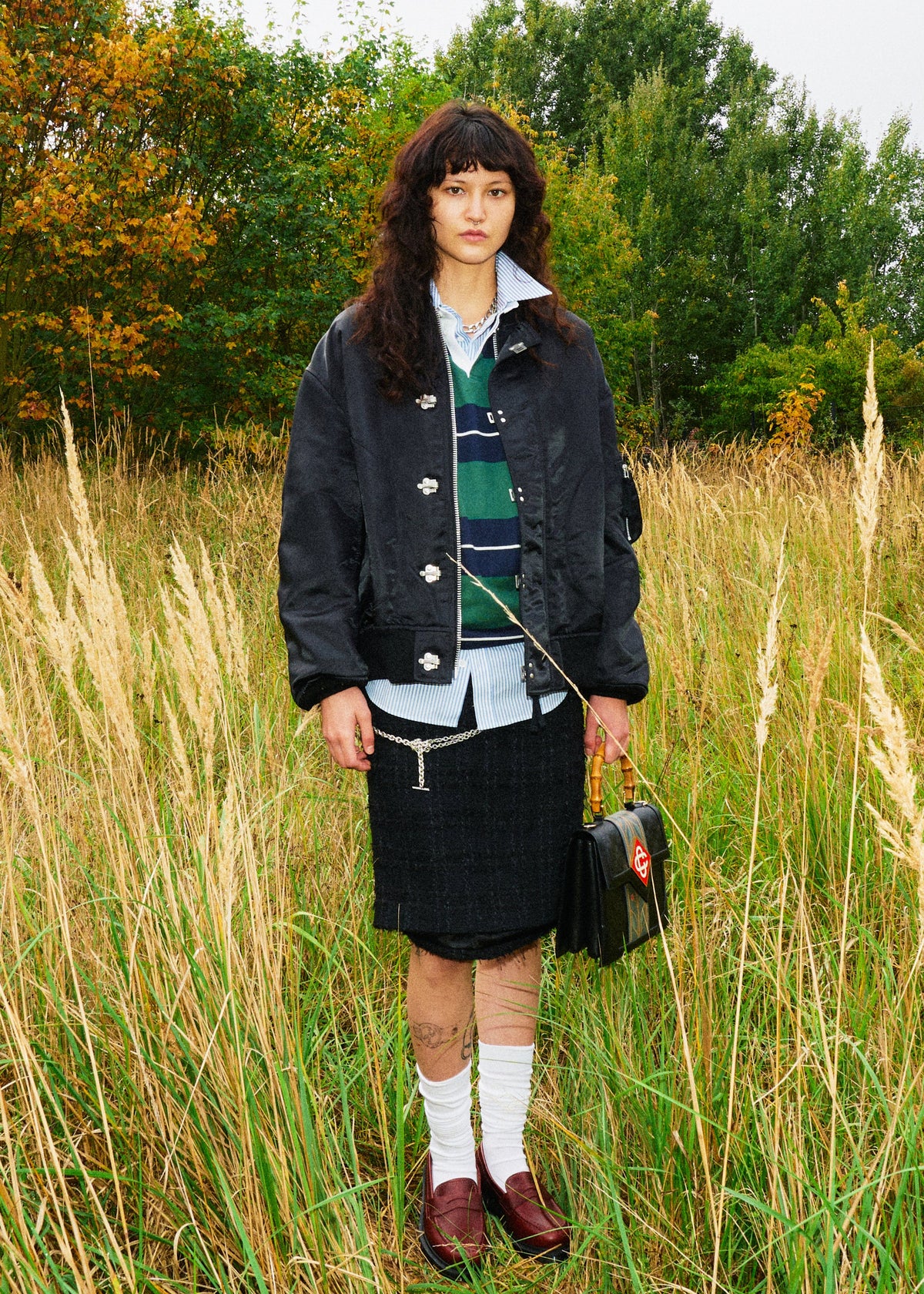 A model stands in a grassy field wearing a dark navy utility jacket over a striped green and navy sweater vest, a white collared shirt, a dark plaid skirt, white socks, and brown loafers, while holding a small black handbag.